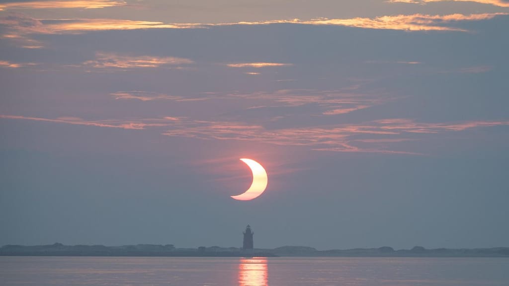 O Sol parcialmente eclipsado a nascer sobre a água, por trás de um farol, com a parte tapada pela Lua bem visível no disco